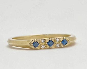 May include: A gold ring with a rectangular design, featuring alternating square blue gemstones and small round clear gemstones. The ring has a smooth, polished finish and is set against a white background.