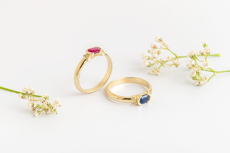 K&ouml;nnte beinhalten: Zwei goldene Ringe mit ovalen Edelsteinen. Der Ring links hat einen roten Rubin, und der Ring rechts hat einen blauen Saphir.