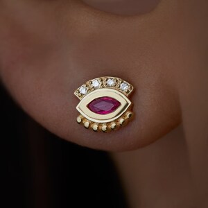 May include: Gold eye-shaped earring with a red gemstone and a row of small white gemstones around the edge.