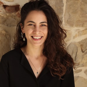 May include: A woman with long, curly brown hair is smiling at the camera. She is wearing a black collared shirt and a silver necklace with a small blue gemstone pendant. She has a gold hoop earring in her left ear and a gold nose ring.