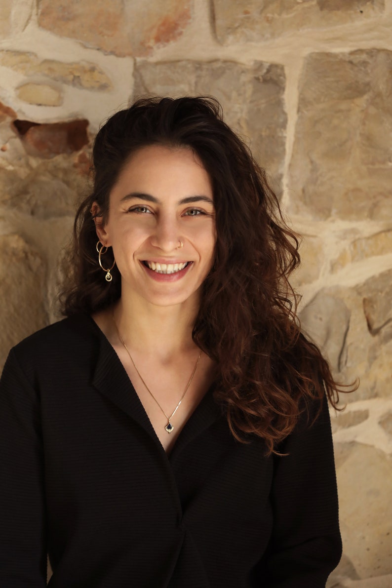 May include: A woman with long, curly brown hair is smiling at the camera. She is wearing a black collared shirt and a silver necklace with a small blue gemstone pendant. She has a gold hoop earring in her left ear and a gold nose ring.