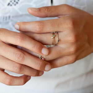 May include: A hand wearing four delicate gold rings with tiny gemstones. The rings feature a green, blue, white, and red stone.