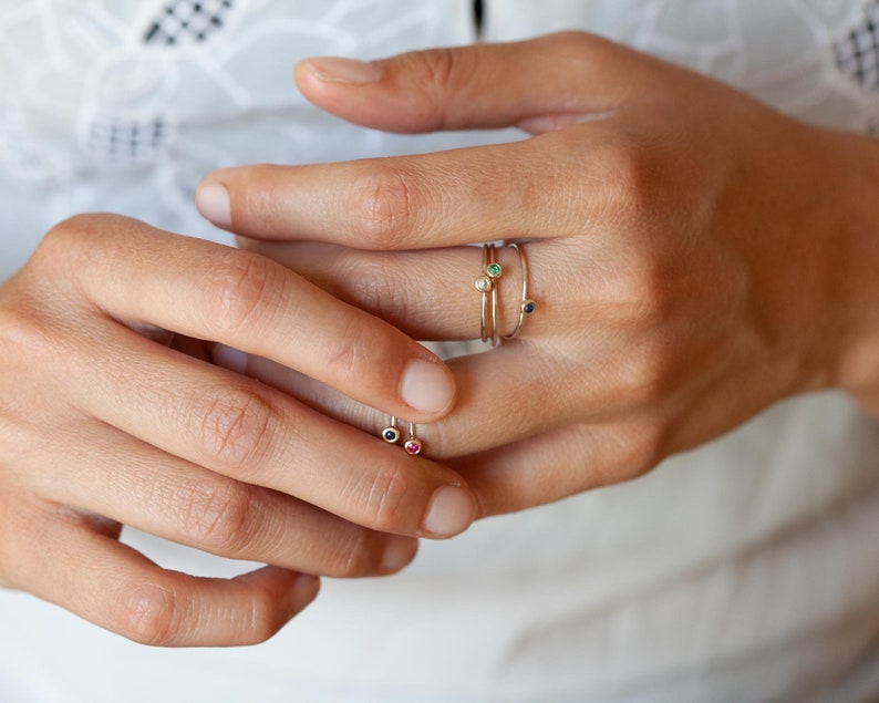 May include: A hand wearing four delicate gold rings with tiny gemstones. The rings feature a green, blue, white, and red stone.