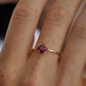 Può includere: Un delicato anello d'oro con un gruppo di quattro pietre preziose rosso scuro disposte a forma di fiore. La sottile fascia lucida dell'anello crea un design minimalista ed elegante. Le pietre preziose sono vicine, formando un piccolo punto focale accattivante.
