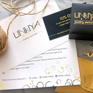 K&ouml;nnte beinhalten: Eine wei&szlig;e Karte mit dem Text "UNKIYA jewelry workshop" und einem 10% Rabatt-Gutscheincode. Die Karte liegt neben einer schwarzen Box mit dem gleichen Text und einem goldenen Schmetterling auf einem gelben Umschlag.
