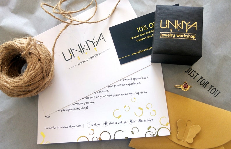 K&ouml;nnte beinhalten: Eine wei&szlig;e Karte mit dem Text "UNKIYA jewelry workshop" und einem 10% Rabatt-Gutscheincode. Die Karte liegt neben einer schwarzen Box mit dem gleichen Text und einem goldenen Schmetterling auf einem gelben Umschlag.