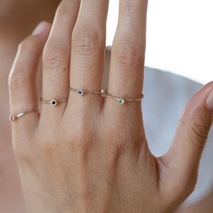 May include: Four delicate gold rings with tiny gemstones. Each ring features a single, small, round gemstone. The gemstones are red, blue, green, and blue.