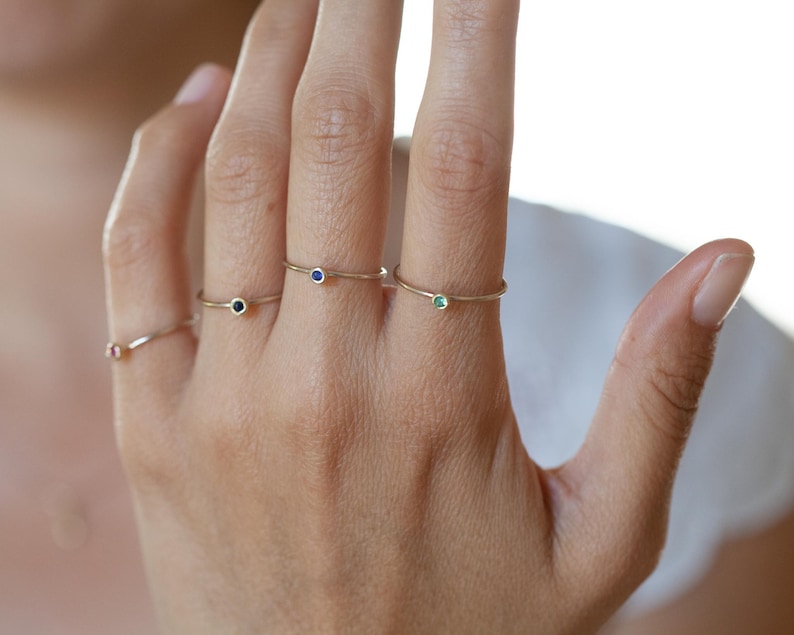 May include: Four delicate gold rings with tiny gemstones. Each ring features a single, small, round gemstone. The gemstones are red, blue, green, and blue.