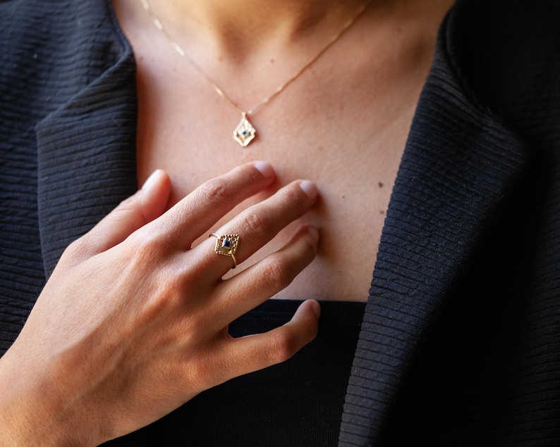May include: A gold ring featuring a blue gemstone set within a geometric design. The ring is worn on the index finger of a person's hand. The person is also wearing a gold necklace with a matching geometric pendant.