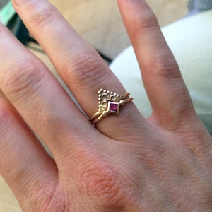 Può includere: Un anello d'oro con una pietra preziosa rosa incastonata in una forma quadrata. L'anello è su un dito con una seconda fascia d'oro.