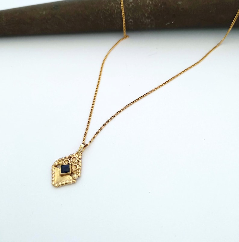May include: A gold chain necklace with a pendant featuring a dark blue square gemstone set in a gold ornate design.