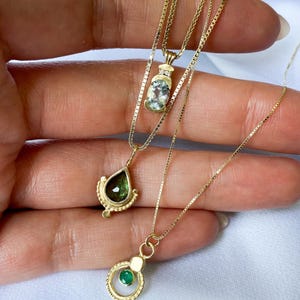 May include: Three gold necklaces with different gemstone pendants. The top necklace has a light blue oval gemstone, the middle necklace has a teardrop-shaped green gemstone, and the bottom necklace has a round green gemstone.