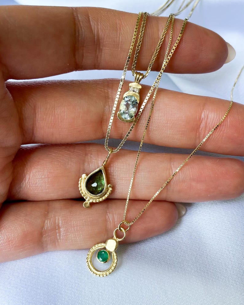 May include: Three gold necklaces with different gemstone pendants. The top necklace has a light blue oval gemstone, the middle necklace has a teardrop-shaped green gemstone, and the bottom necklace has a round green gemstone.