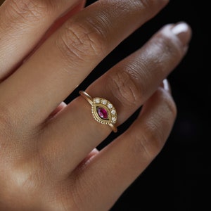 May include: A gold ring with a red gemstone surrounded by smaller white gemstones. The ring is on a person's finger.