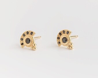 May include: A pair of gold earrings with a crescent moon design. Each earring features a small black gemstone in the center and a row of smaller black gemstones around the edge. The earrings have a textured surface and a cluster of small gold beads hanging from the bottom.
