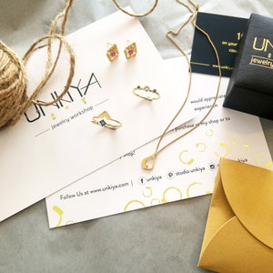 K&ouml;nnte beinhalten: Eine goldene Halskette mit einem Herz-Anh&auml;nger, ein Paar goldene Ohrringe mit rosa und blauen Steinen, ein goldener Ring mit einem blauen Stein und ein goldener Ring mit drei kleinen Steinen. Der Schmuck ist auf einer wei&szlig;en Karte mit dem Text "UNKIYA jewelry workshop" und der Website-Adresse "www.unkiya.com" abgebildet.