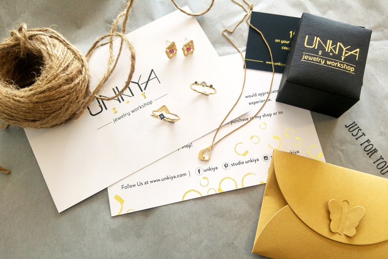 K&ouml;nnte beinhalten: Eine goldene Halskette mit einem Herz-Anh&auml;nger, ein Paar goldene Ohrringe mit rosa und blauen Steinen, ein goldener Ring mit einem blauen Stein und ein goldener Ring mit drei kleinen Steinen. Der Schmuck ist auf einer wei&szlig;en Karte mit dem Text "UNKIYA jewelry workshop" und der Website-Adresse "www.unkiya.com" abgebildet.
