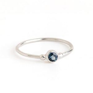 Peut inclure: Une délicate bague en argent avec une petite pierre précieuse bleue ronde sertie au centre. Deux petites perles d'argent sont placées de chaque côté de la pierre précieuse.