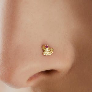 Op de afbeelding: Een gouden neusring met een rode edelsteen in het midden. De ring is waaiervormig en heeft ingewikkelde details.