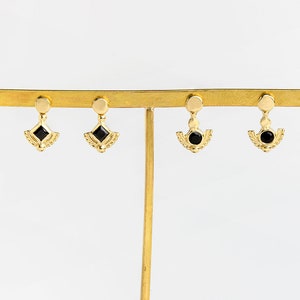 K&ouml;nnte beinhalten: Goldene Ohrringe mit schwarzen Edelstein-Akzenten. Die Ohrringe haben ein zartes, geometrisches Design mit einem herabh&auml;ngenden Tropfen.