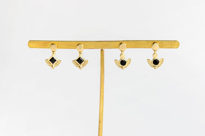 K&ouml;nnte beinhalten: Goldene Ohrringe mit schwarzen Edelstein-Akzenten. Die Ohrringe haben ein zartes, geometrisches Design mit einem herabh&auml;ngenden Tropfen.