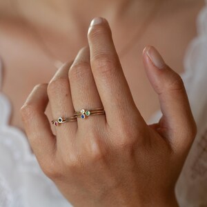 May include: Three gold rings with tiny gemstones. The rings are stacked on a finger. The top ring has a green gemstone, the middle ring has a white gemstone, and the bottom ring has a blue gemstone.
