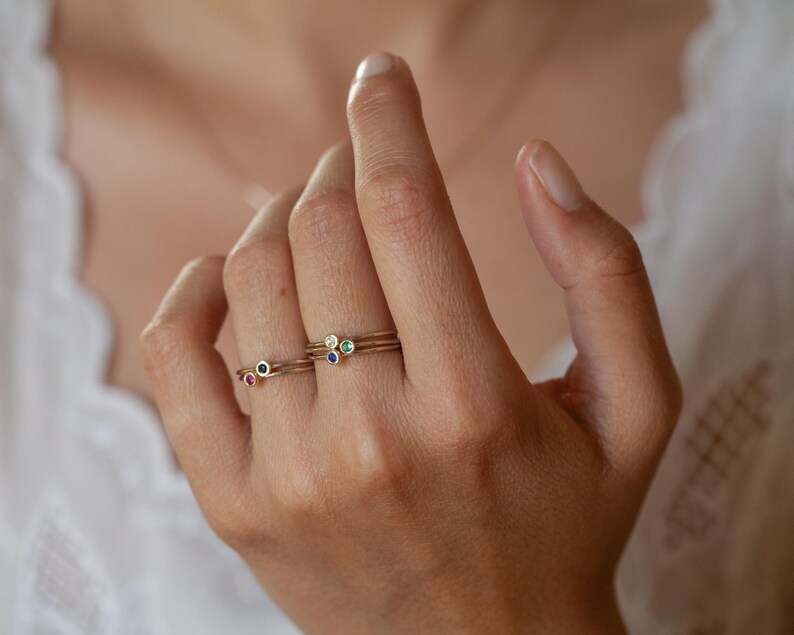 May include: Three gold rings with tiny gemstones. The rings are stacked on a finger. The top ring has a green gemstone, the middle ring has a white gemstone, and the bottom ring has a blue gemstone.