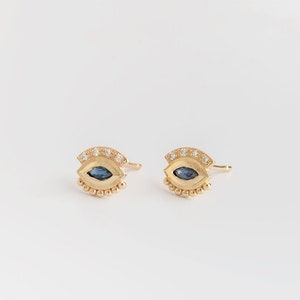 Op de afbeelding: Een paar goudkleurige oorstekers, elk met een oogvormig ontwerp. Het midden van het oog is een donkerblauwe edelsteen, omringd door kleine, heldere edelstenen en een kralenrand. De oorbellen zijn geplaatst tegen een witte achtergrond.
