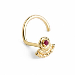 May include: A gold nose stud featuring a curved post and a decorative fan-shaped head. The head is adorned with a small, circular, deep red gemstone set in the center, with a scalloped edge detail along the bottom of the fan-shaped design.