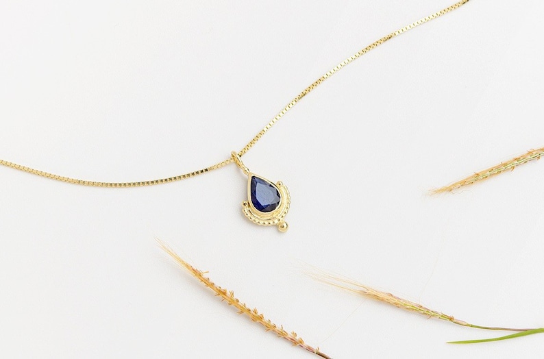 May include: A gold chain necklace with a teardrop-shaped pendant featuring a dark blue gemstone surrounded by a gold filigree design.