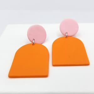 Peut inclure: Une paire de boucles d'oreilles pendantes géométriques orange et rose. Les boucles d'oreilles sont en pâte polymère et présentent une forme d'arche arrondie avec un petit cercle rose sur le dessus.