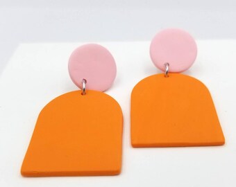 May include: A pair of orange and pink geometric dangle earrings. The earrings are made of polymer clay and feature a rounded arch shape with a small pink circle on top.