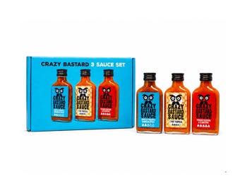 Op de afbeelding: Een Crazy Bastard 3-sausset met een blauwe kartonnen doos en drie glazen flesjes hete saus. De flessen hebben blauwe, gele en rode etiketten, elk met een gestileerd gezichtslogo en de tekst Crazy Bastard Sauce.