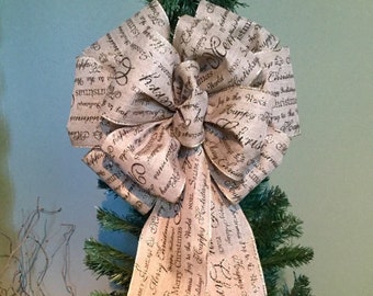 Op de afbeelding: Een kerstboomtopperstrik gemaakt van beige stof met zwart script met de tekst "Vrolijk Kerstfeest" en andere feestelijke groeten. De strik staat op een groene kerstboom.