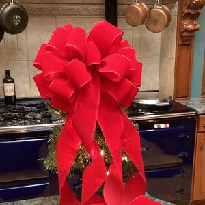May include: A large, red velvet bow with long tails, perfect for a Christmas tree topper. The bow is tied around a small artificial Christmas tree.