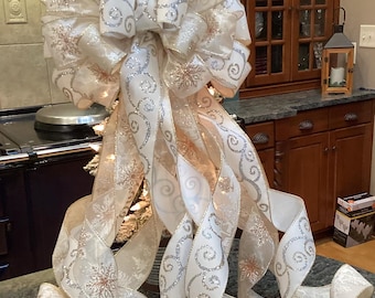 Op de afbeelding: Een grote decoratieve strik gemaakt van wit en champagnekleurig lint met glinsterende zilveren krullen en sneeuwvlokpatronen. De strik heeft meerdere lange, trapsgewijze linten die in lagen naar beneden krullen en draperen.