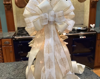 Op de afbeelding: Een grote decoratieve strik gemaakt van wit en goud lint met een herhalend patroon van gouden kerstbomen. Het lint heeft een glinsterende, glitterende textuur en goudkleurige randen, gerangschikt in meerdere lussen en lange, golvende uiteinden.