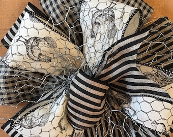 Op de afbeelding: Een decoratieve strik gemaakt van meerdere linten met zwart-wit gingham, zwart-witte strepen en een crèmekleurige stof met een zwarte kip- en gaasprint, geaccentueerd met een laag zilverkleurig metalen kippengaas.