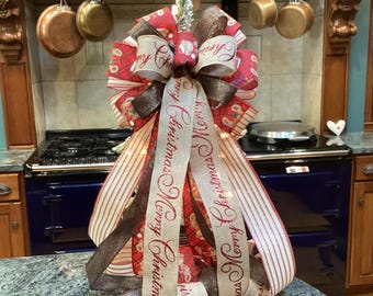 Op de afbeelding: Een decoratieve kerststrik met gelaagde linten in rood, wit en bruin. Het ontwerp bevat ruitpatronen, gestreepte texturen en een lint in jute-stijl bedrukt met de herhalende rode tekst Merry Christmas in een sierlijk lettertype.