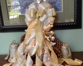 Op de afbeelding: Een decoratieve kerstboomstrik met gouden en zilveren linten. De strik is geplaatst op een kleine kerstboom met lichtjes. De linten vallen naar beneden, aangevuld met dennenappels en decoratieve bellen.
