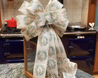 Op de afbeelding: Een decoratieve strik van beige lint met een jute-textuur, voorzien van een herhalend patroon van groene dennenkransen en kleine gouden klokjes. De strik heeft meerdere lussen en lange, afhangende linten die het gedetailleerde motief tonen.