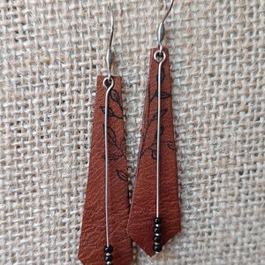 May include: A pair of long, tapered, brown leather earrings featuring a black line-art botanical leaf pattern. Each earring is accented with a vertical silver-toned metal wire and three small black beads at the bottom, suspended from silver-toned hooks.