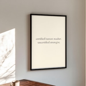 May include: A rectangular wall art print in a thin black frame featuring a cream-colored background with the black text "certified button masher. uncertified strategist." centered in the middle of the frame in a simple serif font.
