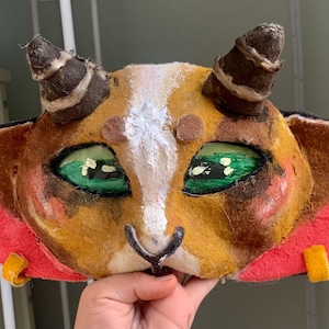 Op de afbeelding: Een handgemaakt wezenmasker met een bruine en beige vachtachtige textuur, twee donkerbruine hoorns en grote, felgroene ogen. Het masker heeft een witte streep in het midden, een zwarte neus en een felrode kraag versierd met gele lipjes.