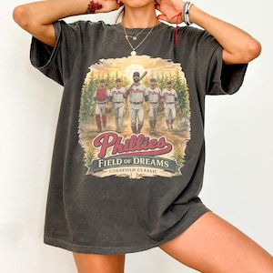May include: A charcoal gray short-sleeve t-shirt featuring a graphic of four baseball players in uniforms standing in a cornfield. The text reads Phillies Field of Dreams Cornfield Classic. The design includes a sunburst background and red lettering.