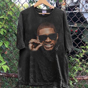 Może przedstawiać: Ciemnoszary t-shirt z krótkim rękawem z dużym nadrukiem graficznym przedstawiającym osobę w czarnych okularach przeciwsłonecznych i srebrnych nakładkach na zęby. Osoba trzyma mały przedmiot przy ustach, a nazwisko Usher jest napisane białą kursywą.