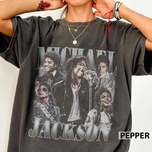 May include: A charcoal gray short-sleeve t-shirt featuring a vintage-style graphic print of Michael Jackson in various poses. The text "MICHAEL JACKSON" is printed in large, bold, distressed lettering across the front of the garment.