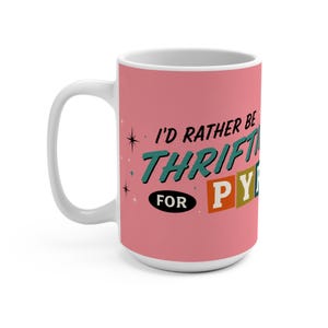 Può includere: Una tazza in ceramica rosa con interno e manico bianchi. L'esterno presenta il testo nero I'D RATHER BE, il testo verde acqua THRIFTING e il testo FOR PYREX con le lettere P, Y, R, E, X all'interno di singoli quadrati arancioni e verde acqua.