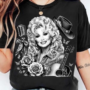 May include: A black short-sleeve t-shirt featuring a detailed black and white graphic print of a woman with voluminous hair, a cowboy hat, a vintage microphone, an acoustic guitar, and several blooming roses and butterflies.
