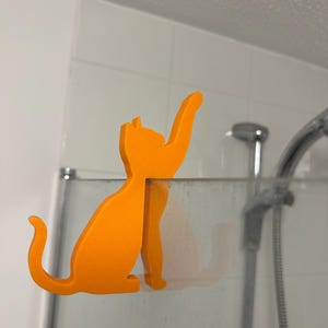 May include: A bright orange, flat, cat-shaped hook designed to hang over the top edge of a glass shower screen. The cat silhouette is depicted in a seated position with one front paw extended upward as if reaching for an object above the glass.